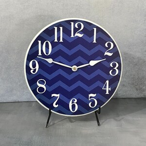 May include: A blue and white chevron patterned clock with white numbers and hands. The clock is on a black stand.