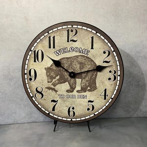 May include: A round, cream-colored clock with black Roman numerals and hands. The clock features a brown bear with a fish in its mouth and the text "Welcome to our den".