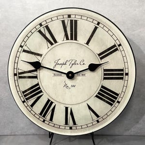 May include: A round wall clock with a distressed off-white face and black Roman numerals. The center features the text Joseph Tyler Co., Boston, Est. 1830, and No. 144. Two black spade-style hands are positioned at the center of the clock face.