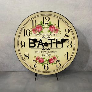 May include: A round, cream-colored clock with black Roman numerals and a black hand. The clock face has a floral design with pink roses and the words "SOAP ON REQUEST", "BATH", "ONE TOWEL ONLY", and "25 Cents".
