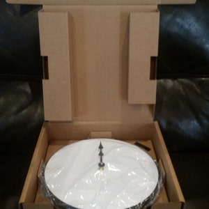 May include: A white clock face with black hands in a cardboard box. The clock face is covered in plastic wrap.