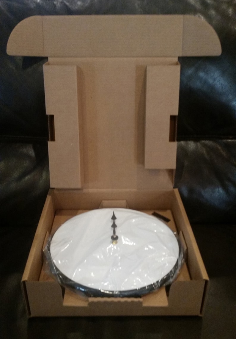 May include: A white clock face with black hands in a cardboard box. The clock face is covered in plastic wrap.