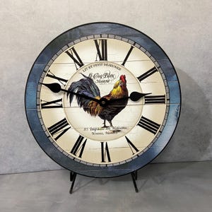 May include: A round, blue and white clock with Roman numerals and a rooster illustration. The clock face features the text "Le Coq Bleu" and "85 Impasse des Maisons, Neuves, Montred".