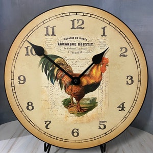 May include: A vintage-style clock with a rooster design. The clock face is cream-colored with black Roman numerals and a black hand. The rooster is in the center of the clock face and is surrounded by a decorative border. The clock face also includes the text "MAGASIN DE MODES LAMARQUE ROUSTIT Rue de la Romane a Fresnoy, A TOULOUSE".