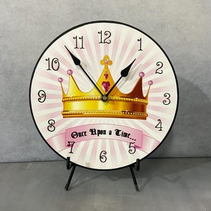 May include: A white clock with a pink and white background. The clock face features a gold crown with pink jewels and black hands. The clock reads "Once Upon a Time..." in a pink banner.