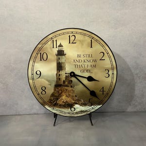 May include: A round wooden clock with a lighthouse image and the text "Be still and know that I am God..." The clock has black hands and black numbers. The clock is on a black stand.
