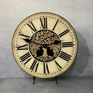 May include: A large, round, cream-colored clock with black Roman numeral markings. The clock face features a black and gold tree of life design. The clock is on a black stand.