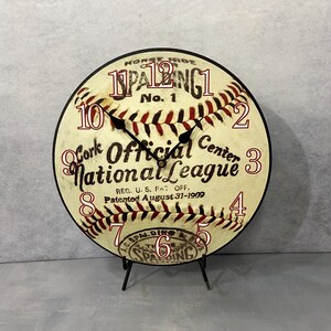 May include: A vintage-style baseball clock with red and white stitching, featuring the text "Horse Hide 11PALBING1 No. 1 Cork Official Center National League REG. U.S. PAT OFF. Patented August 31-1909" and the numbers 1 through 12 around the edge.