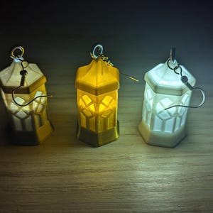 May include: Three small, decorative lantern earrings. The lanterns are made of white, yellow, and clear plastic. Each lantern has a small, clear window on each side. The earrings are hanging from silver hooks.