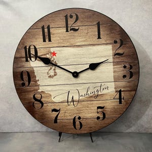 May include: A wooden clock with a rustic brown finish features a white outline of the state of Washington with a red star marking the location of Seattle. The clock face has black Roman numeral hour markers and black hands. The words "Washington" are written in cursive on the clock face.