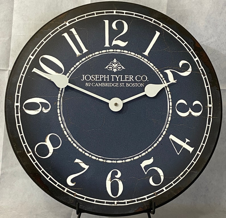May include: A black and white clock with a blue face. The clock has Roman numerals and a decorative design around the edge. The clock face says "Joseph Tyler Co. 82 Cambridge St. Boston".