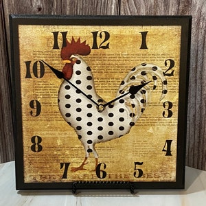 May include: A black framed square clock with a white rooster with black polka dots on a vintage-style background. The clock face has black hands and black Roman numeral numbers.