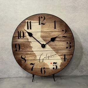 May include: A round wooden clock with a brown wood grain background. The clock face features a white outline of California, black numerals, and black clock hands. The word "California" is written in cursive, with a red star near the number 5.