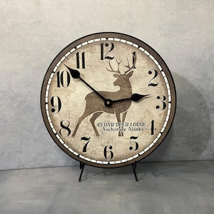May include: A round, wooden clock with a black frame and a distressed white face. The clock features a silhouette of a deer with antlers and the text "CEDAR DEER LODGE Anchorage Alaska".