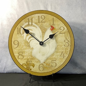 May include: A round, cream-colored clock with a rooster design and black hands. The clock face has a distressed, vintage look with black Roman numerals and a white rooster with a red comb. The clock is on a black stand.