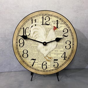 May include: A white rooster clock with black hands and Roman numerals on a vintage newspaper print background. The clock is on a black stand.