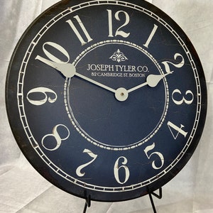May include: A black and white clock with a distressed finish. The clock face is navy blue with white numbers and a white border. The clock is on a black stand. The clock face reads "JOSEPH TYLER CO. 82 CAMBRIDGE ST. BOSTON".