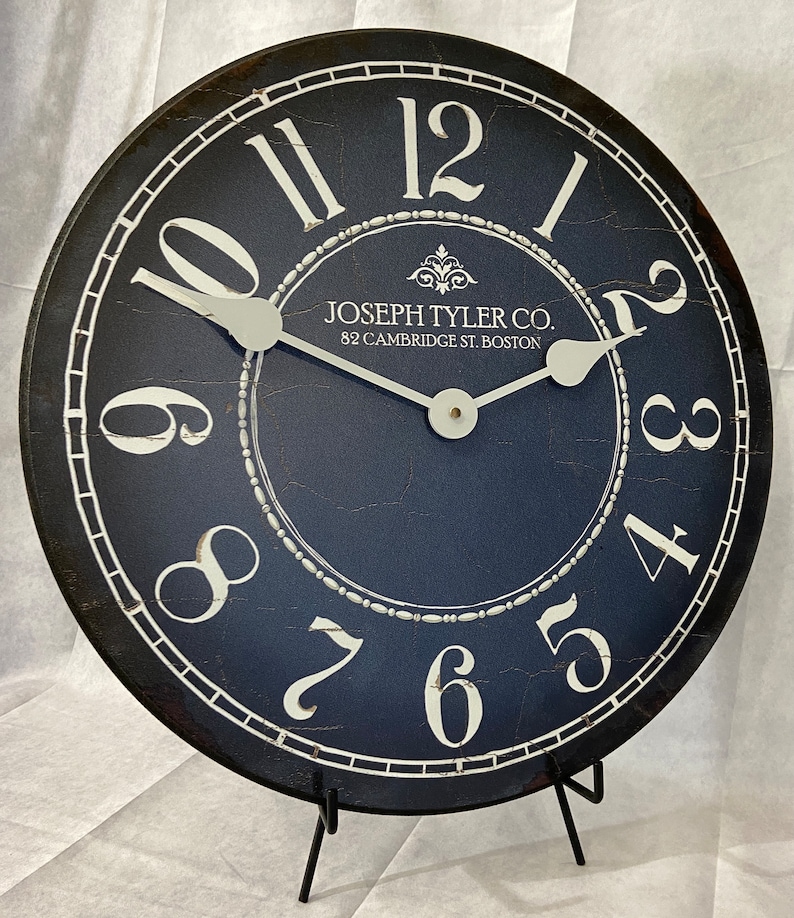 May include: A black and white clock with a distressed finish. The clock face is navy blue with white numbers and a white border. The clock is on a black stand. The clock face reads "JOSEPH TYLER CO. 82 CAMBRIDGE ST. BOSTON".