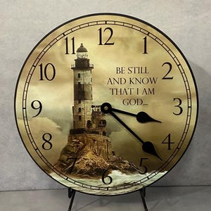 May include: A round wall clock featuring a sepia-toned image of a lighthouse on a rocky shore with crashing waves. The clock face displays black Arabic numerals and the text BE STILL AND KNOW THAT I AM GOD.... It has two black hands.