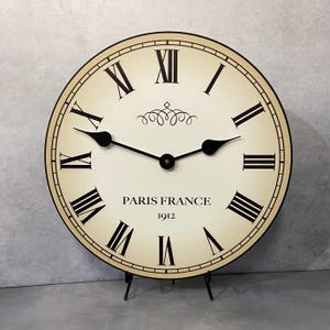 May include: A large, round, cream-colored clock with black Roman numeral markings and black hands. The clock face reads "Paris France 1912". The clock is on a black stand.