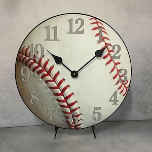 May include: A white baseball-themed clock with red stitching and black hands. The clock face has black Roman numeral hour markers and is set on a black stand.