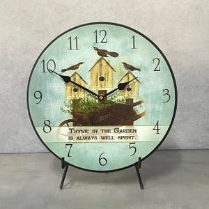 May include: A round, blue and green clock with a black frame and black hands. The clock face features a whimsical design with three birdhouses, birds, and a wheelbarrow with the text "THYME IN THE GARDEN IS ALWAYS WELL SPENT."
