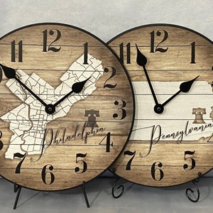 May include: Two wooden clocks with black hands and numbers. The clock on the left features a map of Philadelphia with the Liberty Bell icon. The clock on the right features a map of Pennsylvania with the Liberty Bell icon. Both clocks have a rustic wood grain finish.