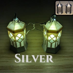 May include: A pair of silver earrings shaped like small lanterns with a white and gold design. The lanterns are lit up with a warm white light.