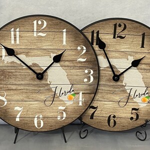 May include: Two wooden clocks with a rustic wood grain design. The clocks feature a white outline of the state of Florida with an orange graphic. The clocks have black hands and white numbers. The text "Florida" is written in cursive on each clock.