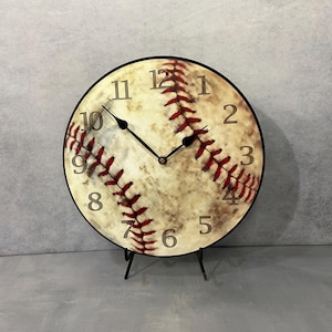 May include: A round, black-framed clock with a baseball design. The clock face is a vintage-style baseball with red stitching and a white background. The numbers are in a brown font.
