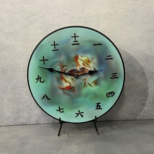 May include: A round, teal-colored clock with black Roman numerals and a black clock face. The clock face features two gold fish swimming in a blue and green watercolor-like background. The clock is on a black stand.