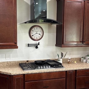 May include: A kitchen scene featuring a brown-framed clock with black hands and numbers, mounted on a white wall. A black range hood is above a gas stovetop with a pan and griddle. Dark brown cabinets are on either side.