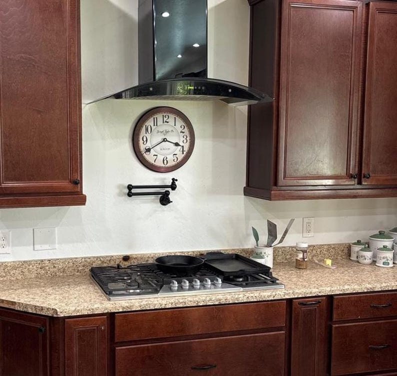 May include: A kitchen scene featuring a brown-framed clock with black hands and numbers, mounted on a white wall. A black range hood is above a gas stovetop with a pan and griddle. Dark brown cabinets are on either side.