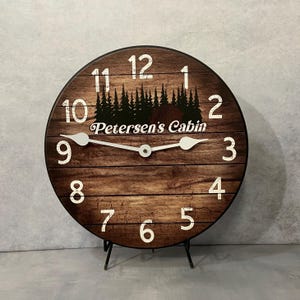 May include: A brown wooden clock with white numbers and hands. The clock has a black silhouette of trees and the text "Petersen's Cabin" on the face.