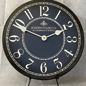 May include: A black and white clock with Roman numerals and a distressed blue face. The clock is on a black stand and has the text "JOSEPH TYLER CO. 82 CAMBRIDGE ST. BOSTON" on the face.