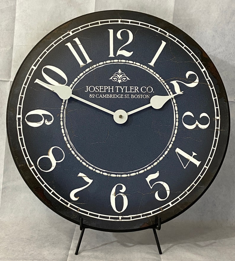 May include: A black and white clock with Roman numerals and a distressed blue face. The clock is on a black stand and has the text "JOSEPH TYLER CO. 82 CAMBRIDGE ST. BOSTON" on the face.