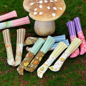 Pode incluir: Uma coleção de meias de boneca coloridas com vários padrões, incluindo listras, desenhos florais e formas geométricas. As meias estão dispostas em uma superfície verde gramada com um cogumelo decorativo no fundo.