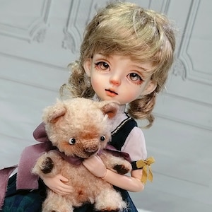 May include: A doll with blonde hair wearing a blue and green plaid dress with a white lace trim and holding a brown teddy bear. The doll is sitting on a white pedestal.