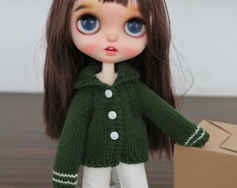 May include: A green knitted cardigan with white trim, white trousers, and white trainers. The doll is holding two brown cardboard boxes.