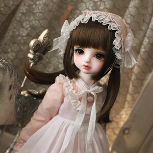 May include: A porcelain doll with brown hair, wearing a pink and white dress with lace trim and a white lace headband. The doll has green eyes and a pink blush on her cheeks.