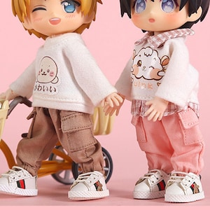 May include: Two dolls, one with blonde hair and a white shirt with a cartoon seal, and the other with black hair and a white shirt with a cartoon sheep, are standing next to a yellow toy tricycle. The doll with blonde hair is wearing brown pants and the doll with black hair is wearing pink pants. Both dolls are wearing shoes.