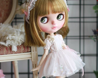 May include: A doll with long blonde hair wearing a white dress with lace details and a pink headband. The doll is standing on a white surface with white flowers.