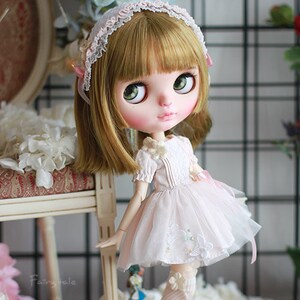 May include: A doll with long blonde hair wearing a white dress with lace details and a pink headband. The doll is standing on a white surface with white flowers.