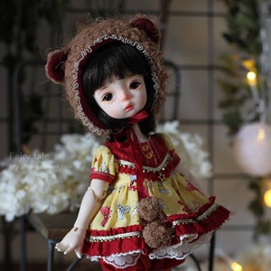 May include: A small doll with brown hair wearing a yellow and red dress with a brown bear hat. The doll is standing on a wooden surface with white flowers in the foreground.