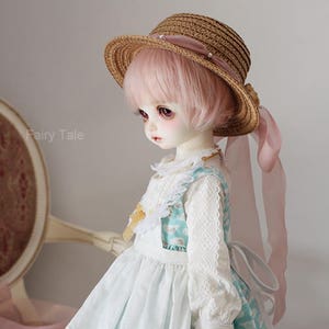 May include: A doll wearing a white dress with lace trim and a blue and white patterned pinafore. The doll has pink hair and is wearing a brown straw hat with a pink ribbon.