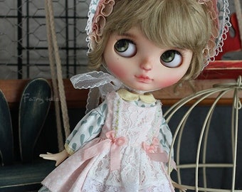May include: A doll wearing a pink and green dress with a white lace overlay, a yellow hat with flowers, and yellow socks with white polka dots. The doll is standing on a wooden floor with a red wagon and a birdcage in the background.