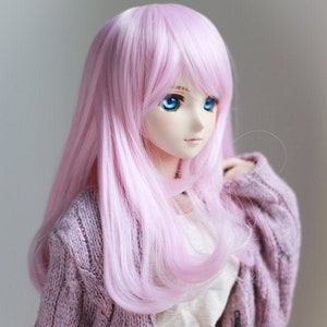 Puede incluir: Una muñeca rosa con el pelo largo, liso y rosa y ojos azules. La muñeca lleva un cárdigan rosa claro sobre una camisa blanca y una falda rosa. La muñeca mira hacia la derecha.