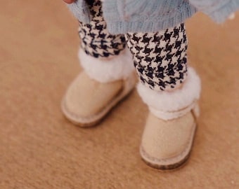 May include: A doll wearing a light blue cardigan with brown buttons, black and white houndstooth pants, a beige scarf, and beige boots. The doll is standing on a wooden surface. The text "SISQ DESIGN FOR DOLLS" is visible in the lower right corner of the image.