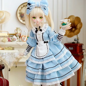May include: A doll wearing a blue and white dress with a black bow and white apron. The doll has blonde hair and green eyes. The doll is holding a teacup and saucer. The doll is standing on a wooden base. The doll is surrounded by furniture and a cat.