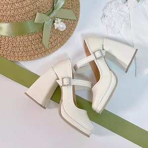 May include: A pair of white heeled shoes with a buckle detail. The shoes are on a white surface with green ribbon.